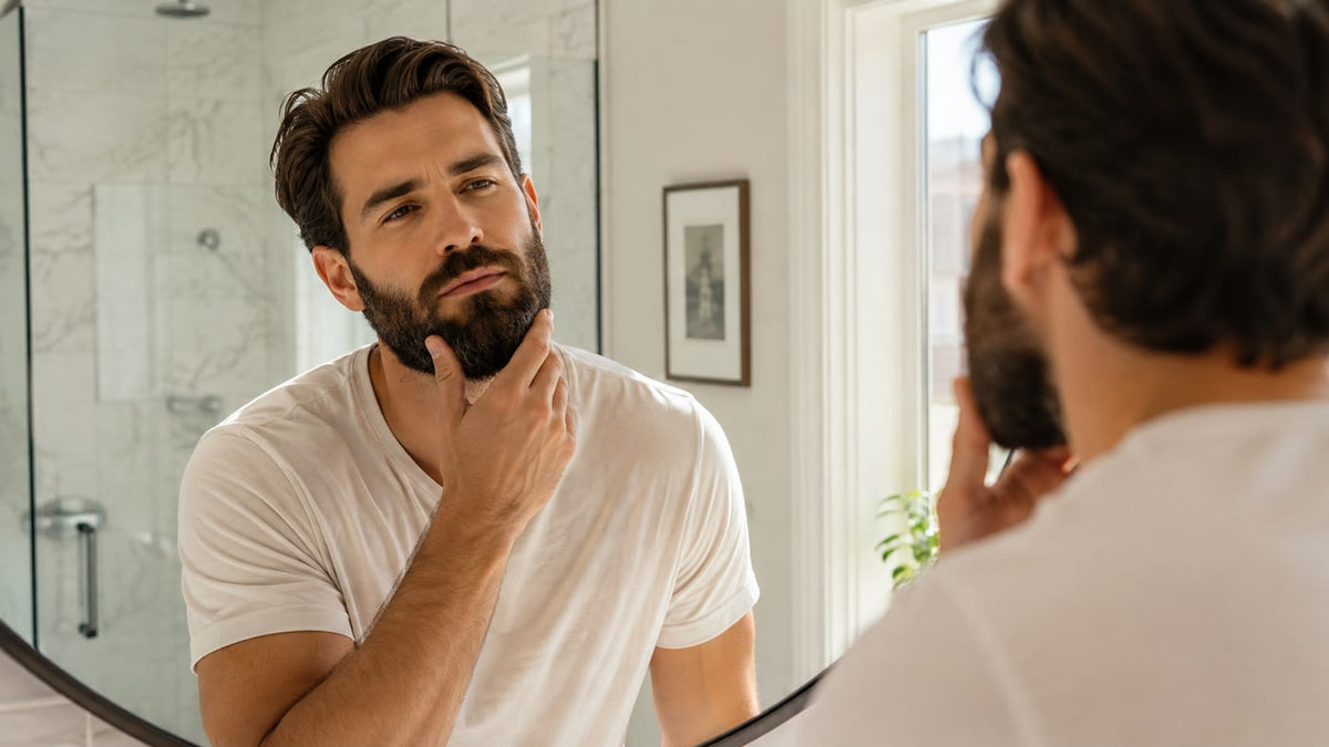 Man with a full groomed beard checking his reflection in a bright modern bathroom mirror
