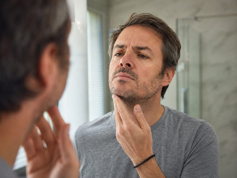Man with short uneven facial hair checking early beard grow-out in a bathroom mirror