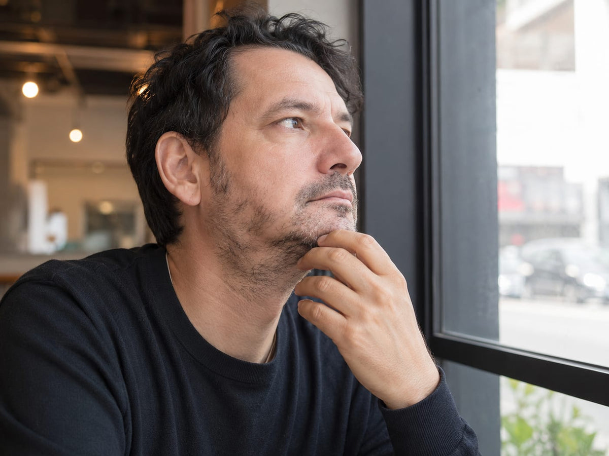 Man with thin patchy stubble looking out a cafe window, hand under his chin, black sweater