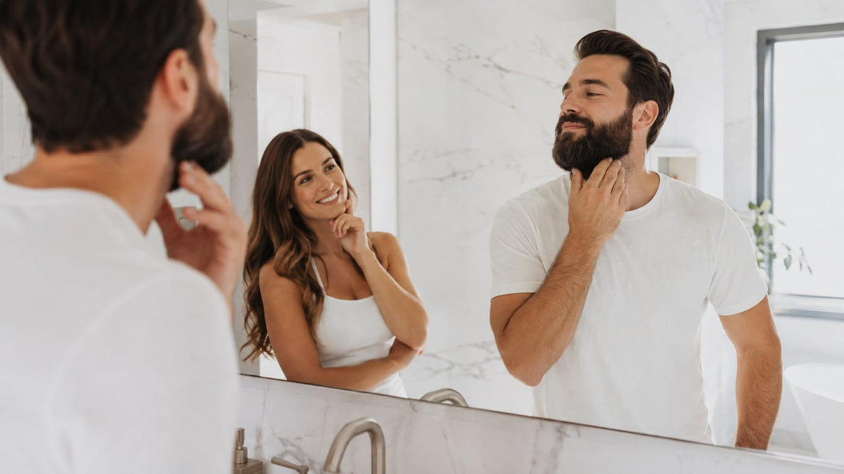 Man with a full dark beard checking his jawline in a bright bathroom mirror